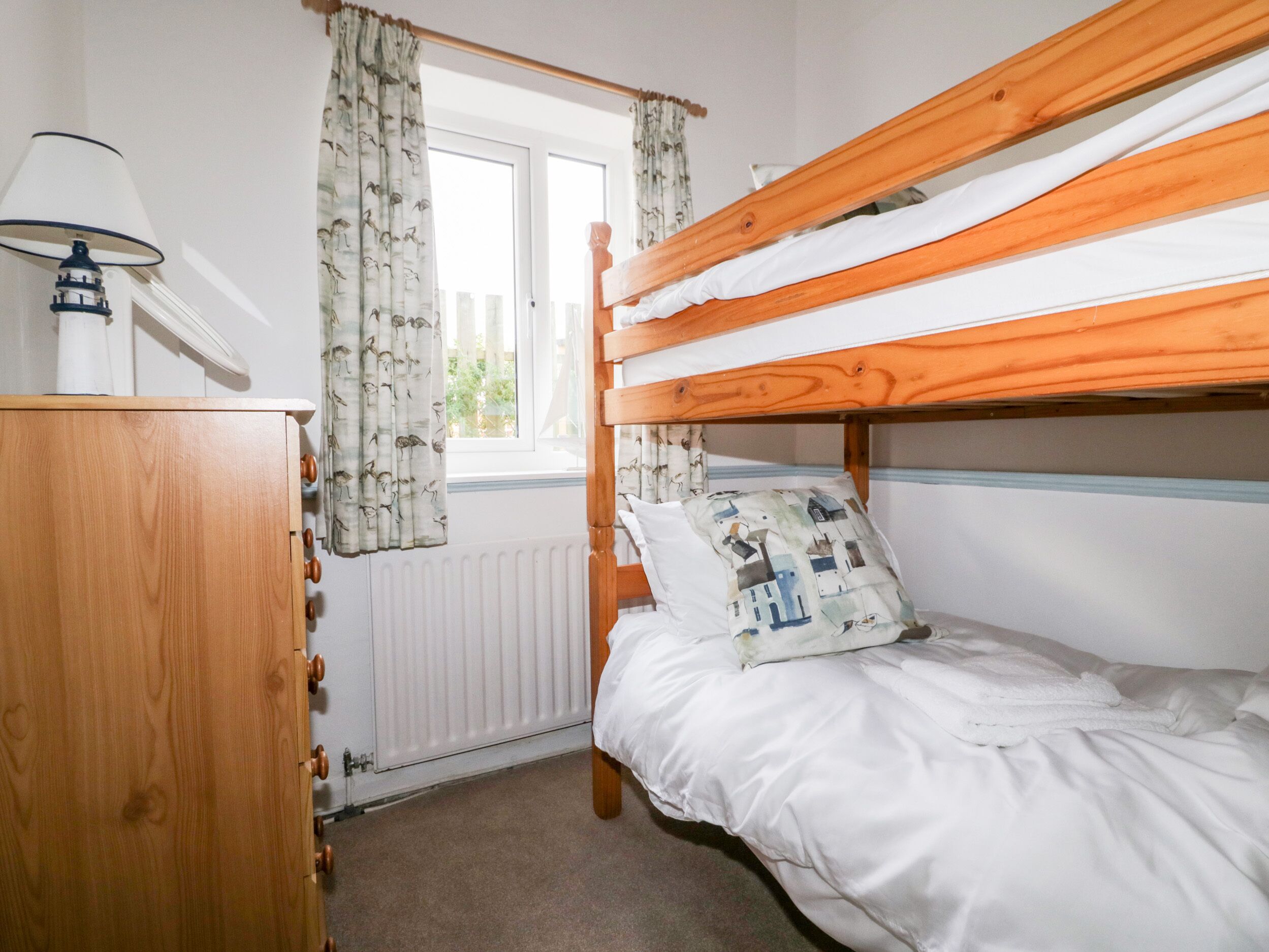 A bunk bed with white bedding, a wooden dresser, and a window with curtains.