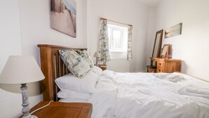 A neatly made bed with white linens, a wooden nightstand with a lamp, a wooden dresser with a mirror, and a window with patterned curtains. - Gull Cottage (Tintagel)