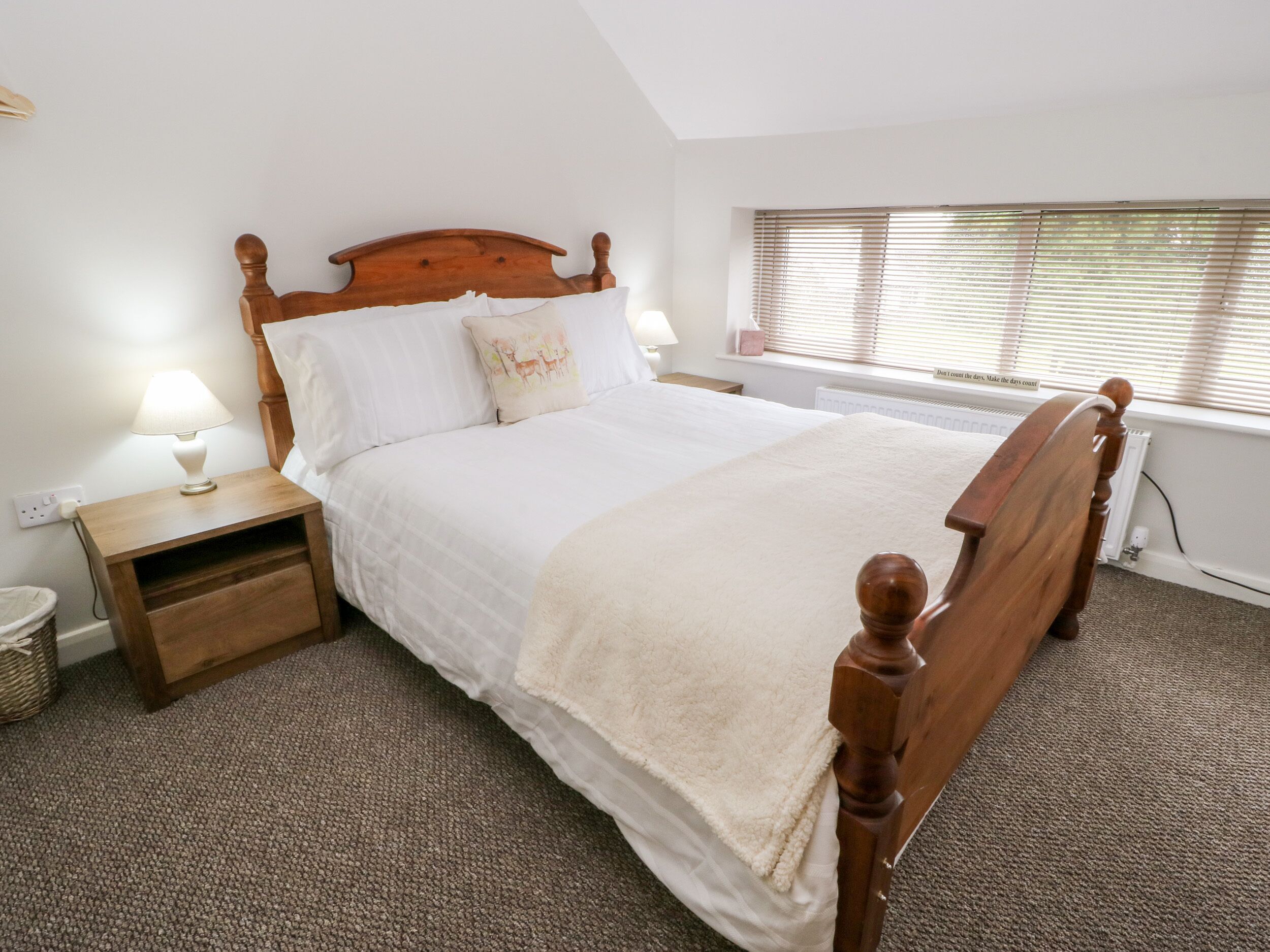 A neatly made bed with a wooden headboard, a bedside table with a lamp, and a window with blinds.