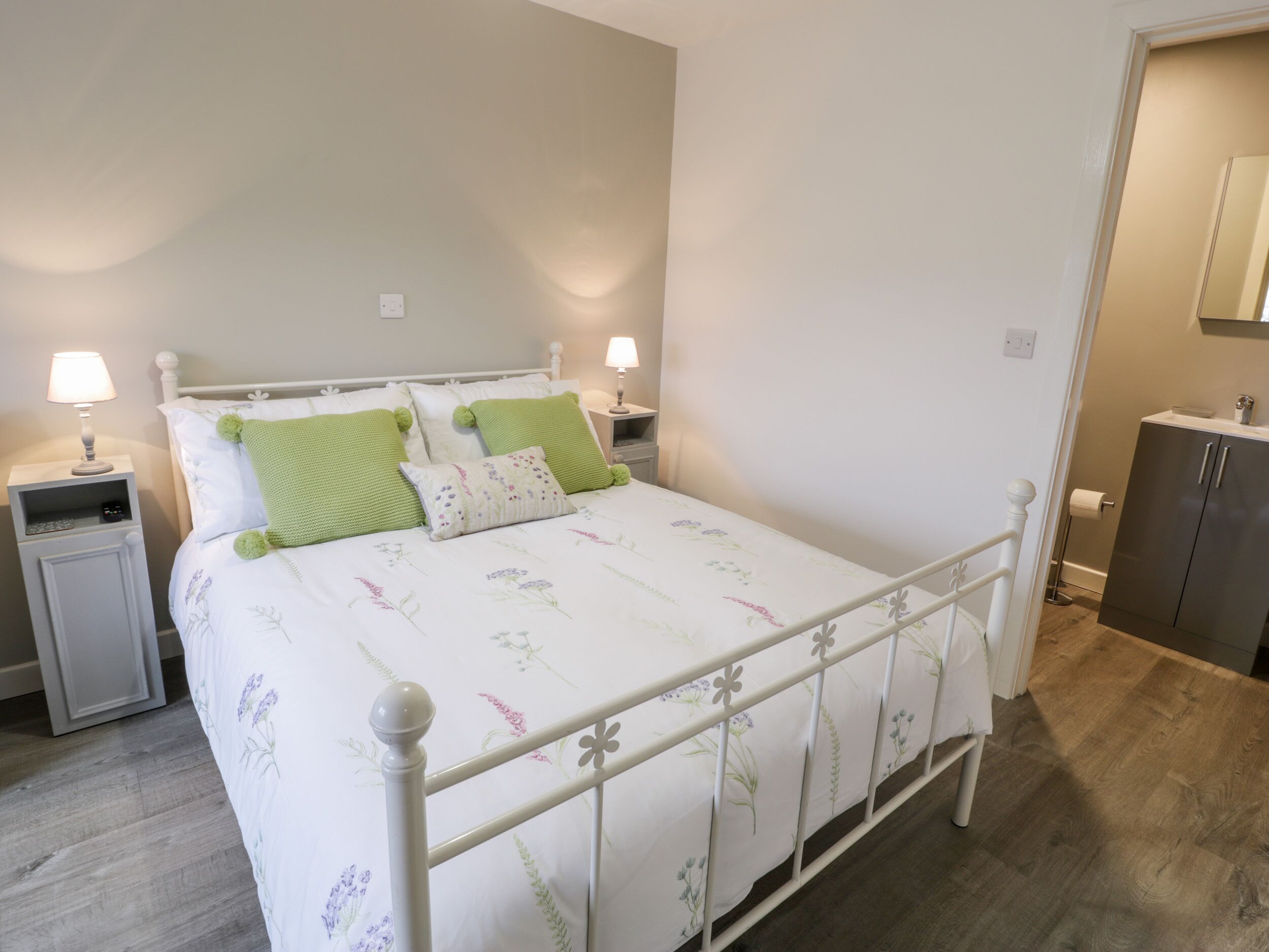 Een netjes ingerichte slaapkamer met een bed met een wit metalen frame, een beddengoed met bloemenprint en groene kussens. Er staan nachtkastjes met lampen en een klein kastje met een spiegel.