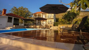 Una piscina con sillas de descanso y sombrillas, una casa de techo rojo y una colina al fondo.