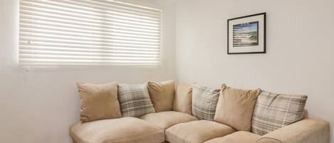 A beige sectional sofa with plaid cushions, a framed picture on the wall, and a window with blinds.