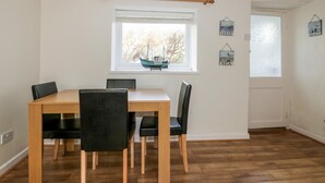 A dining area with a wooden table and four chairs, a window with a view of trees, and a door with a frosted glass panel. - Cove Cottage (Tintagel)