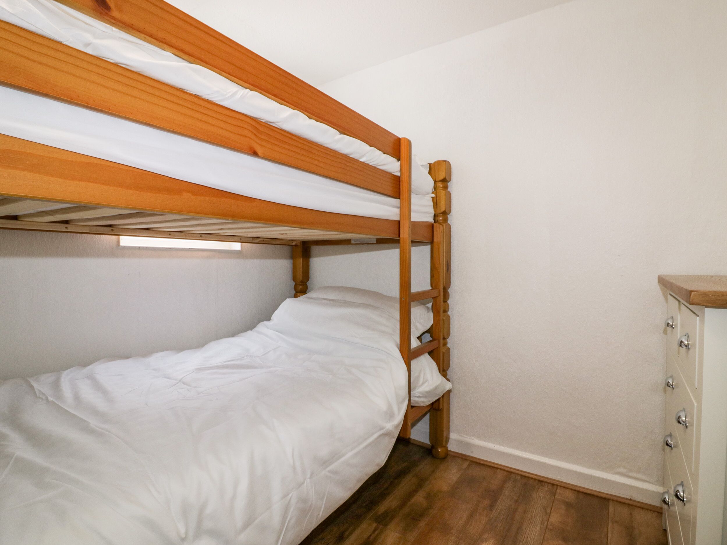 Una cama individual en literas con ropa de cama blanca, cabecera de madera y un pequeño aparador con cajones.