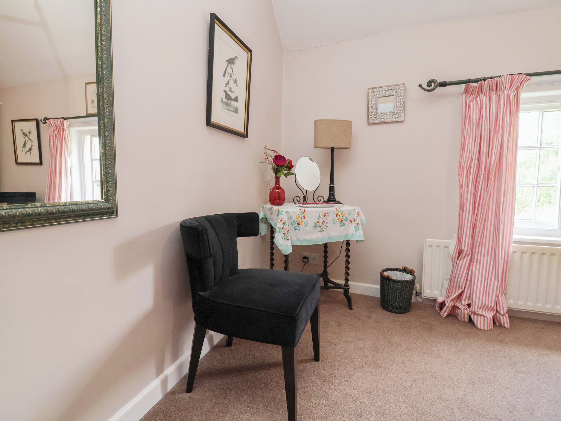 A room with a black chair, a small table with a mirror, a vase with flowers, a radiator, and a window with curtains.