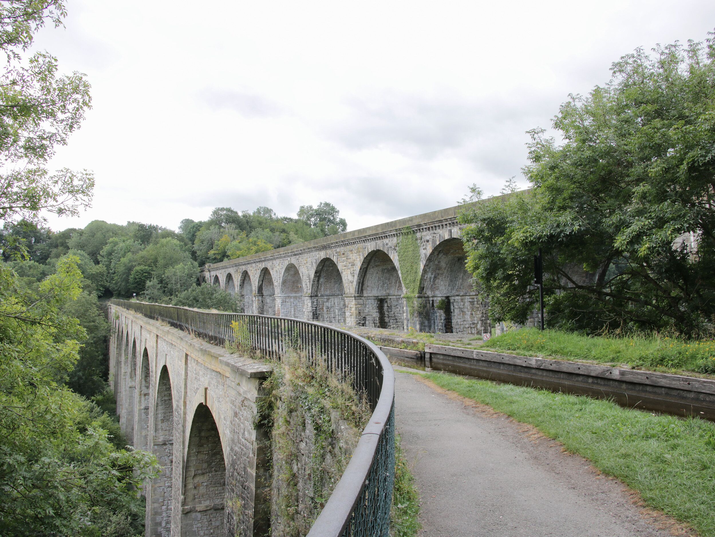 A stone viaduct with multiple arches, surrounded by lush greenery and a paved path.