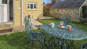 A patio with a round metal table and chairs, a small garden, and a stone building in the background.