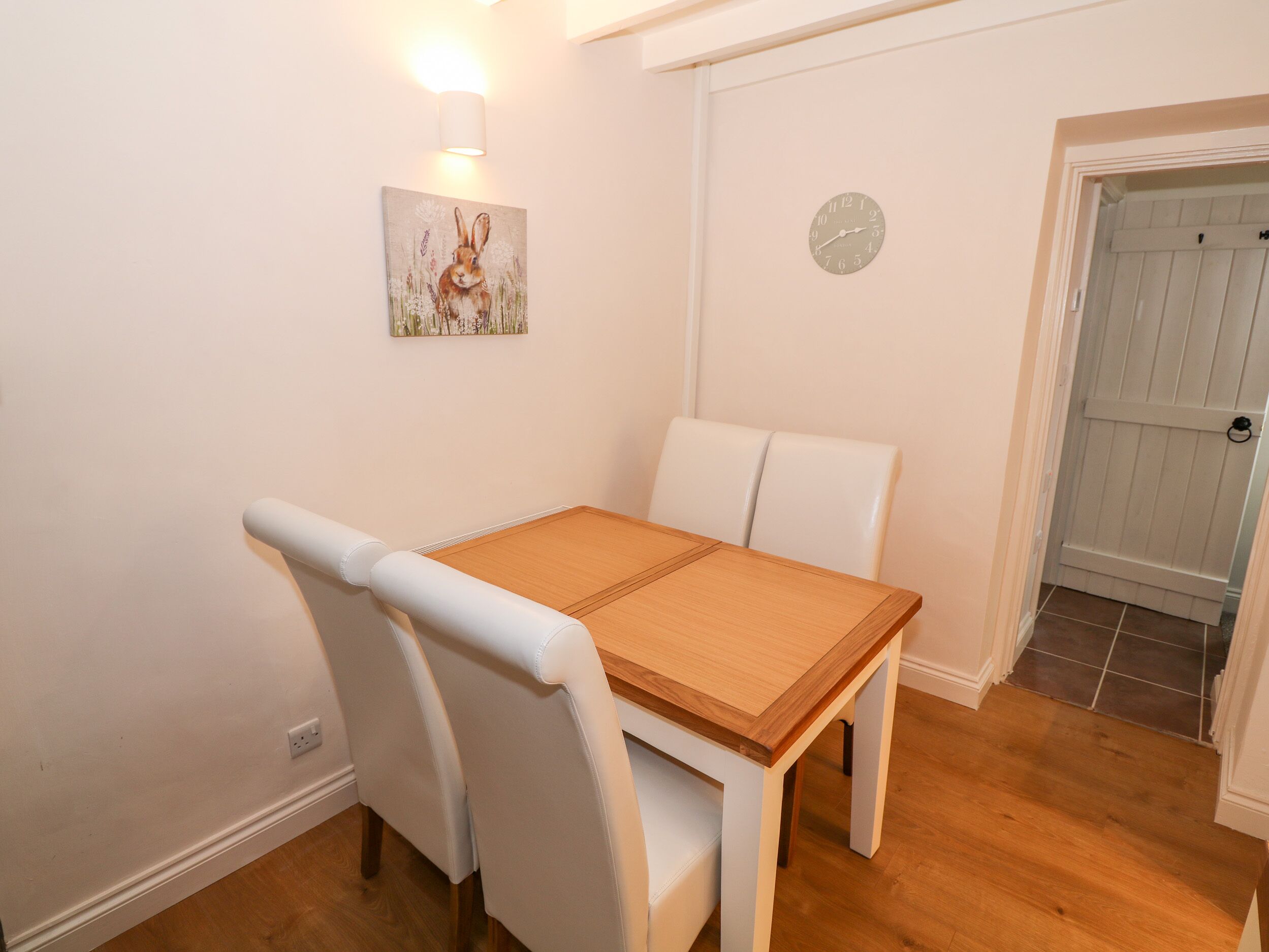 A dining area with a wooden table and white chairs, a wall clock, and a painting of a rabbit.