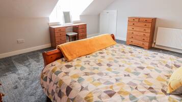 A bedroom with a sloped ceiling, a bed with a patterned bedspread, a wooden dresser, a desk with a chair, and a window providing natural light.