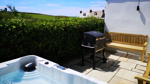 A hot tub, a barbecue grill, and a wooden bench on a patio with a white wall and a hedge in the background. - Trevena Lodge (Tintagel)