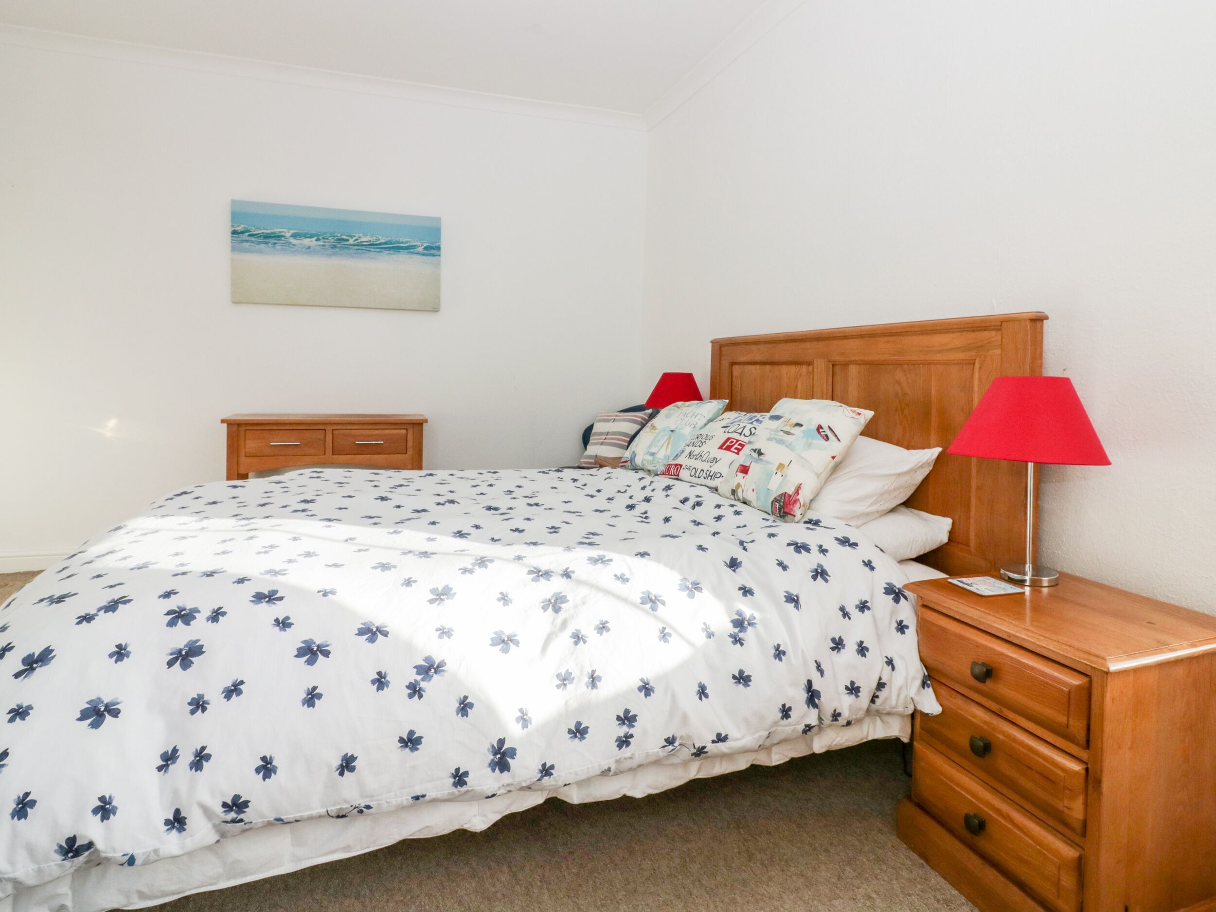 A neatly made bed with a floral bedspread, two bedside tables with lamps, a small bedside table, and a wall-mounted picture of a beach scene.