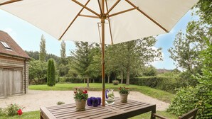 Ein Holztisch mit Stühlen steht unter einem großen Sonnenschirm, umgeben von einem Garten mit Topfpflanzen und einem Kiesweg.