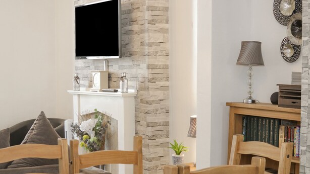 Een woonkamer met een open haard, een aan de muur bevestigde televisie, een boekenplank en een eethoek met houten stoelen.