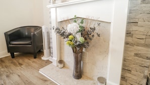 Een woonkamer met een witte open haard, een bloemenvaas, een klok en een zwarte leren fauteuil.