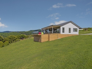 Ein modernes Haus mit einer Holzterrasse, großen Fenstern und Blick auf einen grünen Hügel.