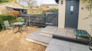 Une maison en pierre avec une porte sombre, un petit patio avec une table et des chaises, et une clôture en bois.