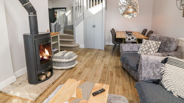 Un salon chaleureux avec un poêle à bois, un escalier, un coin repas et un canapé gris agrémenté de coussins à motifs.