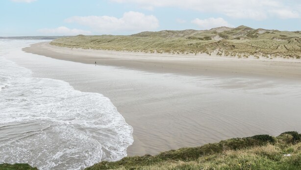 En strand med vågor, en person som går och sanddyner i bakgrunden.