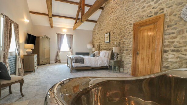 A bedroom with a stone wall, a wooden door, a bed, a nightstand, a TV, and a large bathtub. - Skyfell (Skipton)