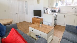 Un espacio habitable compacto con sofá, mesa de centro, televisor, zona de cocina con microondas, horno y fregadero, además de un reloj en la pared.