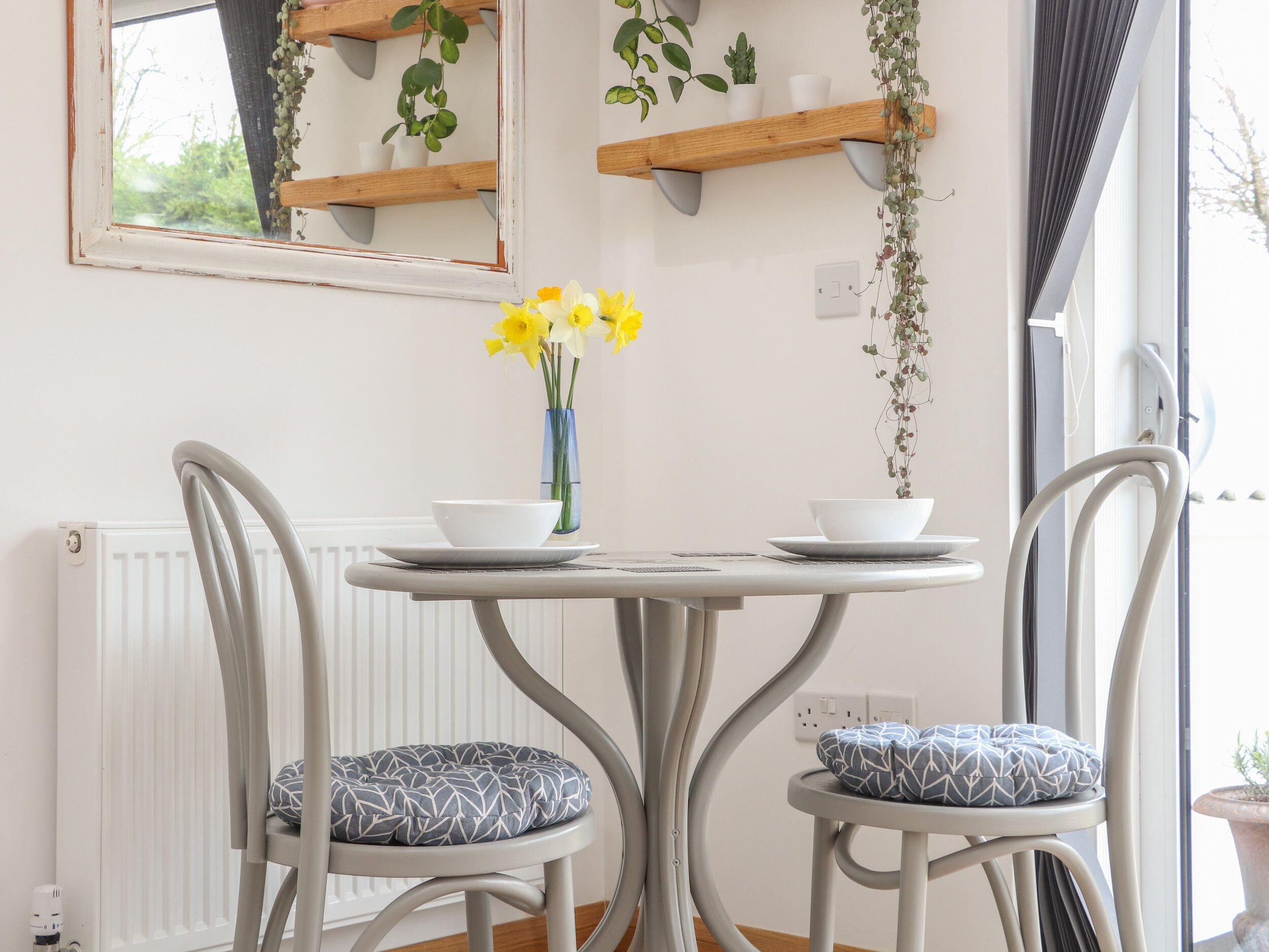 Uma área de refeições com uma mesa redonda, duas cadeiras com almofadas estampadas, um vaso com flores amarelas e prateleiras de madeira com plantas.