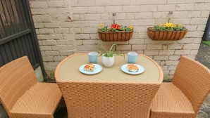 A wicker table with two chairs, two plates with food, two cups, and two hanging flower baskets against a brick wall. - Hazelmere Cottage (Worksop)