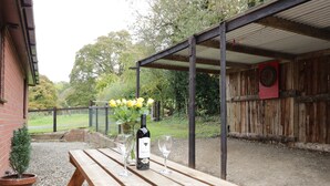 A wooden picnic table with a bottle of wine and glasses, set outdoors with a brick wall and a wooden fence in the background. - The Alders (Llanfyllin)