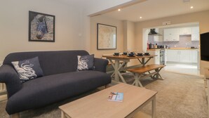 A living room with a dark blue sofa, a wooden coffee table, and a dining table with chairs. There is a kitchen visible in the background.