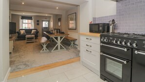 A modern kitchen with a gas stove, microwave, and white cabinetry. There is a dining area with a table and chairs, and a living room with a sofa and a television.