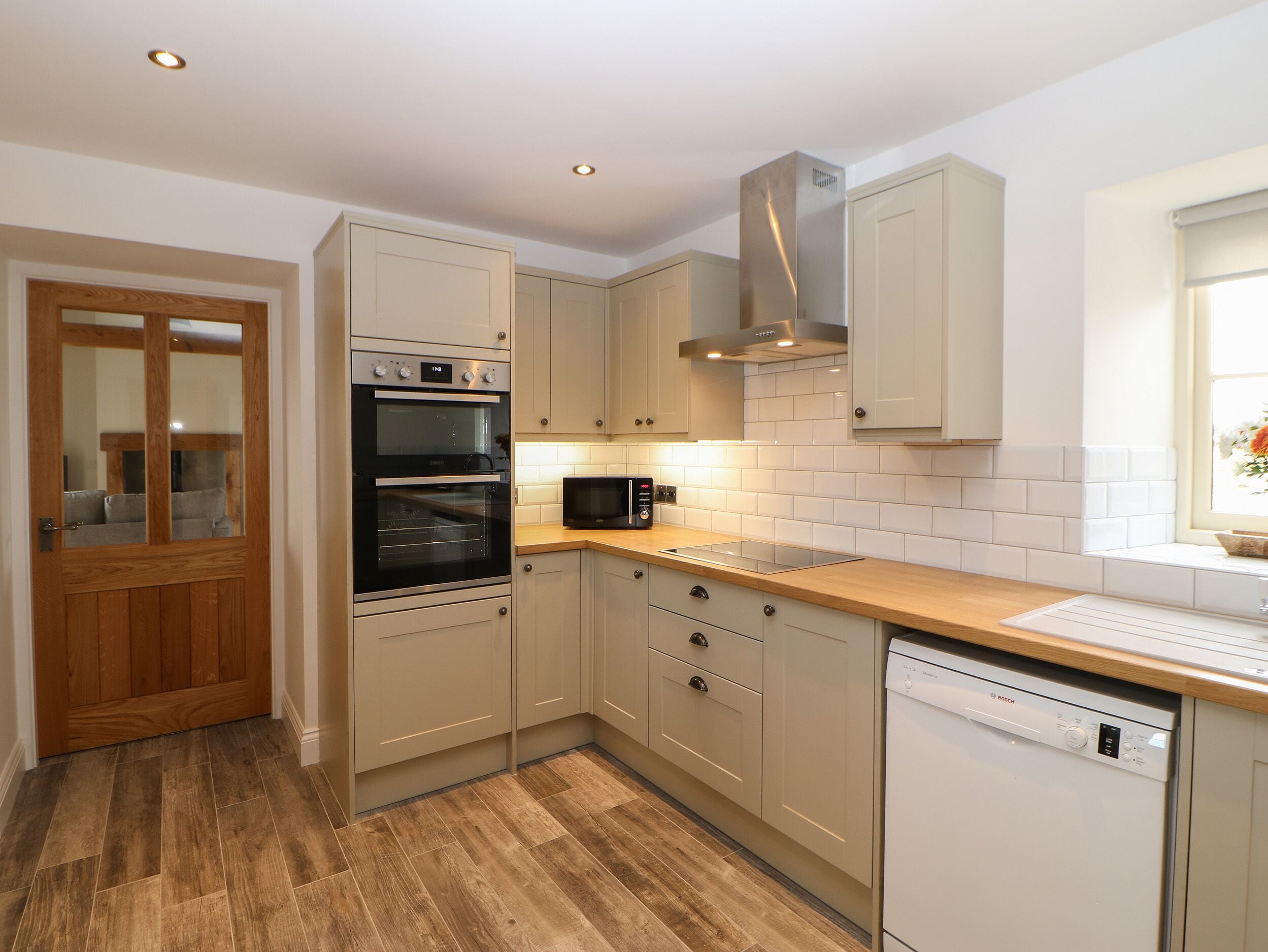 A modern kitchen with wooden flooring, beige cabinetry, a built-in oven, a microwave, and a Bosch dishwasher.