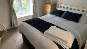 En snyggt bäddad säng med vita och mörkblå kuddar, ett träbord vid sängen med en vit lykta och ett fönster med gardiner.