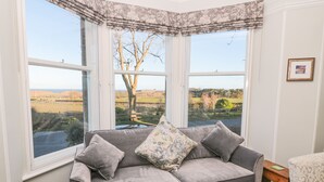 A corner room with a grey sofa, two cushions, and a view of a rural landscape through large windows. - Whinstones at 4 The Villas (Alnwick)