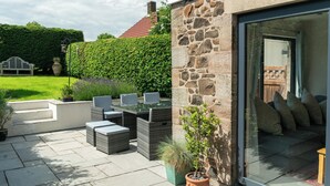 A patio with outdoor furniture, a stone wall, and a glass door leading to a room with a sofa and cushions. - Whinstones at 4 The Villas (Alnwick)
