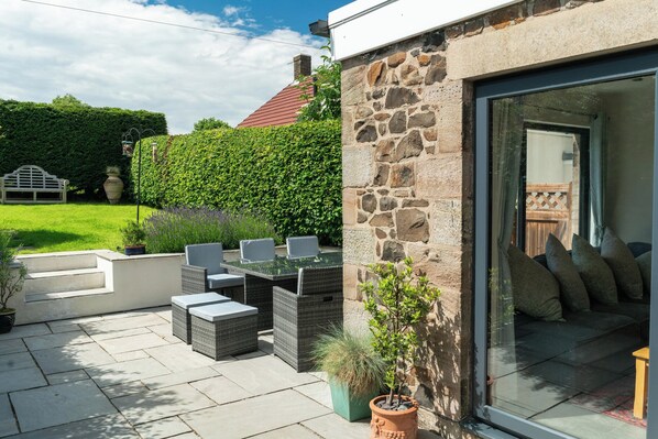 A patio with outdoor furniture, a stone wall, and a glass door leading to a room with a sofa and cushions.