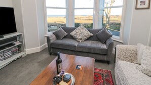 A living room with a grey sofa, a wooden coffee table with a bottle of wine, a plate of cheese and grapes, and a red rug. - Whinstones at 4 The Villas (Alnwick)