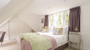 A bedroom with a sloped ceiling, a bed with multiple pillows, a nightstand with a lamp, and a window with curtains. - The Lodge (Norwich)