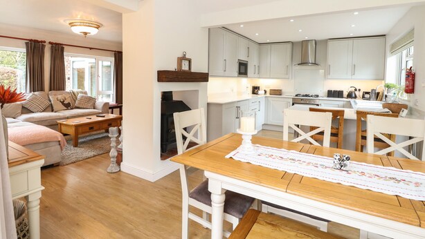 A modern kitchen with white cabinets, a dining table with a floral table runner, and a cozy living area with a sofa and a fireplace. - The Lodge (Norwich)