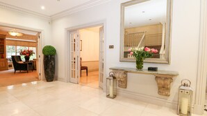 A spacious hallway with a marble floor, a large mirror, a console table with a vase of flowers, and a potted plant. - Gleneagles (Ascot)