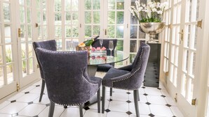 A dining area with a glass table, two velvet chairs, a champagne bottle, and a vase of flowers. - Gleneagles (Ascot)