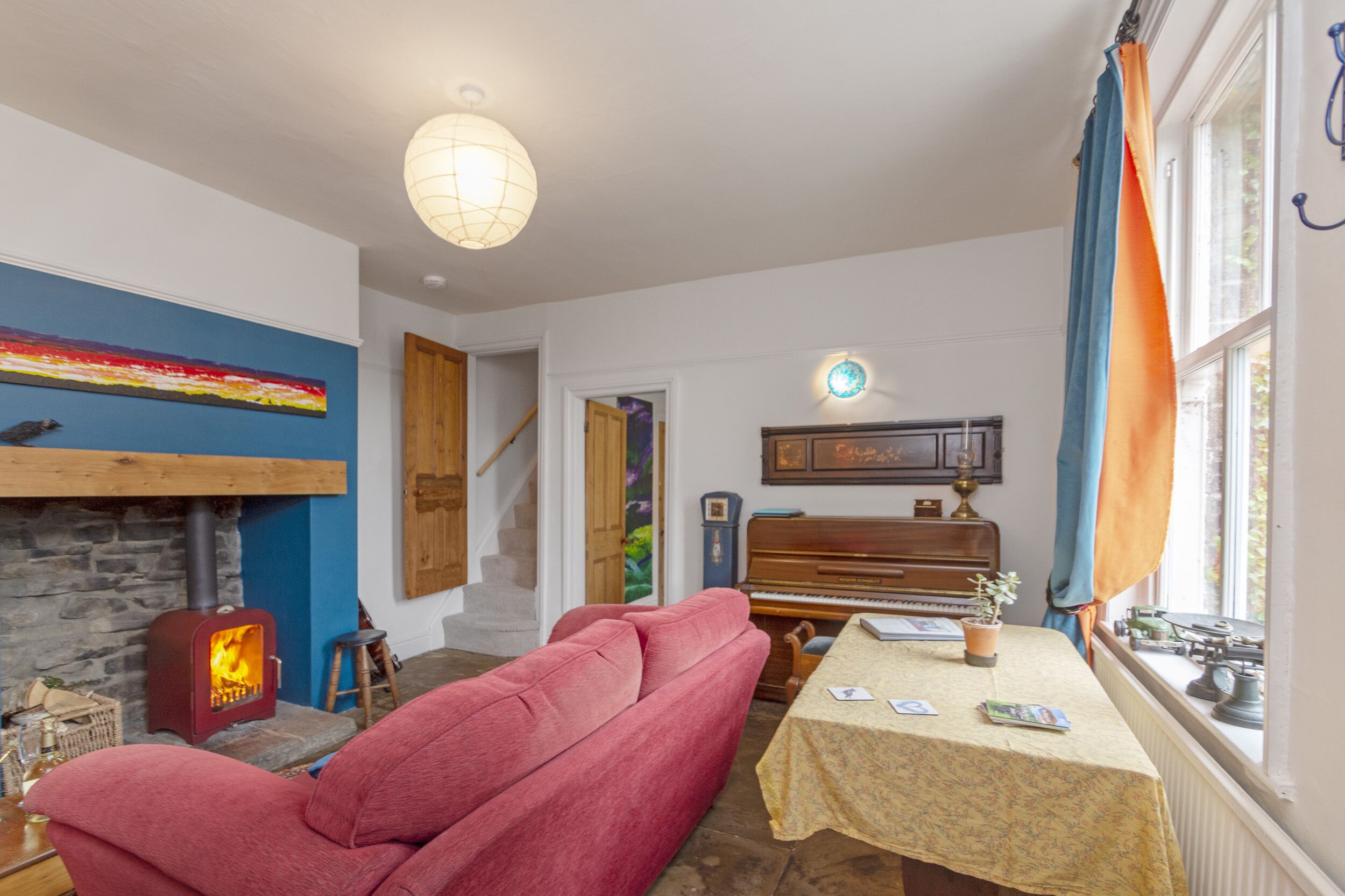 A cozy living room with a stone fireplace, a red sofa, a piano, and a table with books and a lamp.