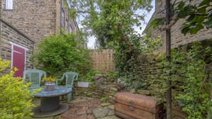A stone-paved courtyard with a rustic wooden chest, green chairs, and a table with a vase of flowers. - Rose Cottage (Keighley)