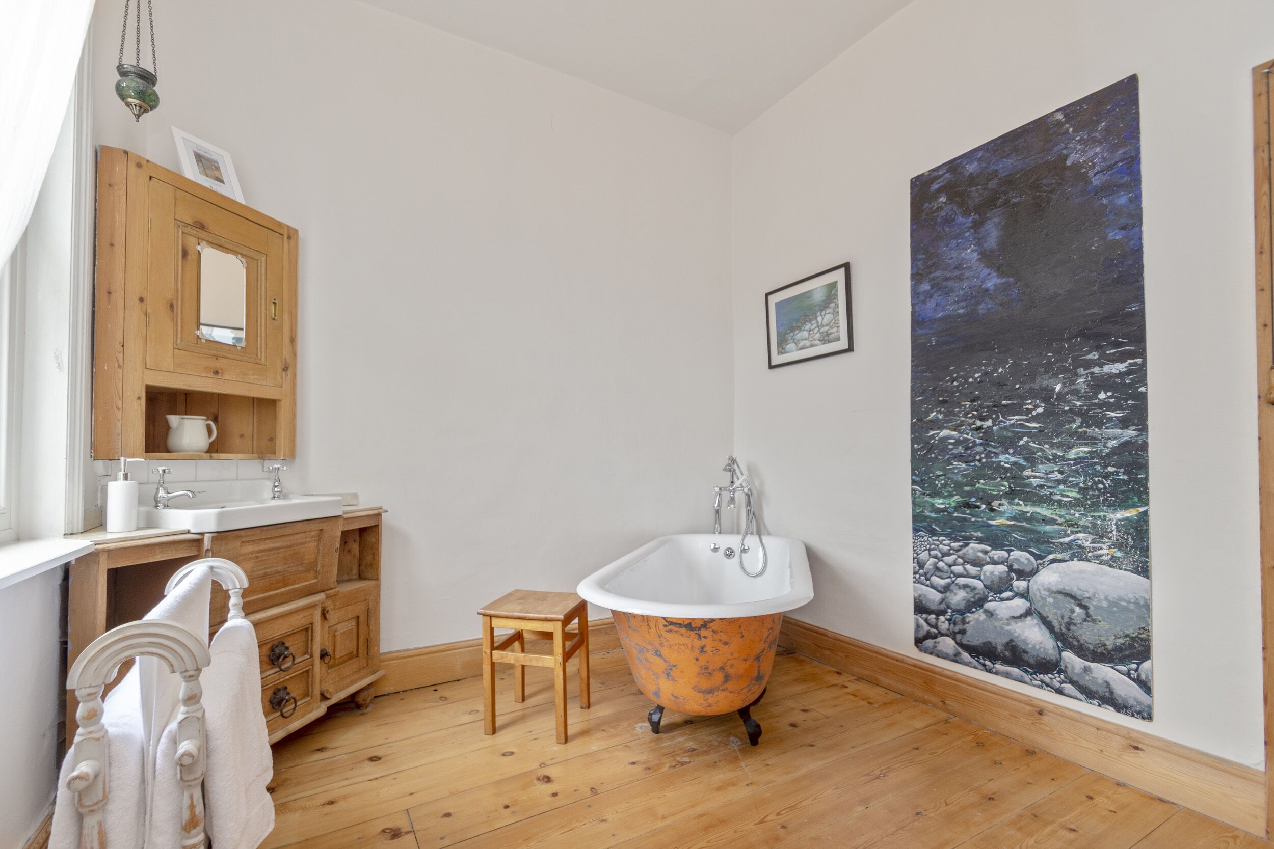 A bathroom with a wooden vanity, a white sink, a freestanding bathtub, a small wooden stool, and a large wall mural.