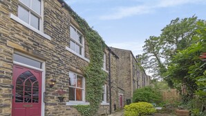 A stone building with a red door and windows, surrounded by greenery and a stone pathway. - Rose Cottage (Keighley)