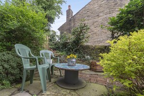 A stone building with a chimney, a round table with a flower pot, and two green chairs in a garden area. - Rose Cottage (Keighley)