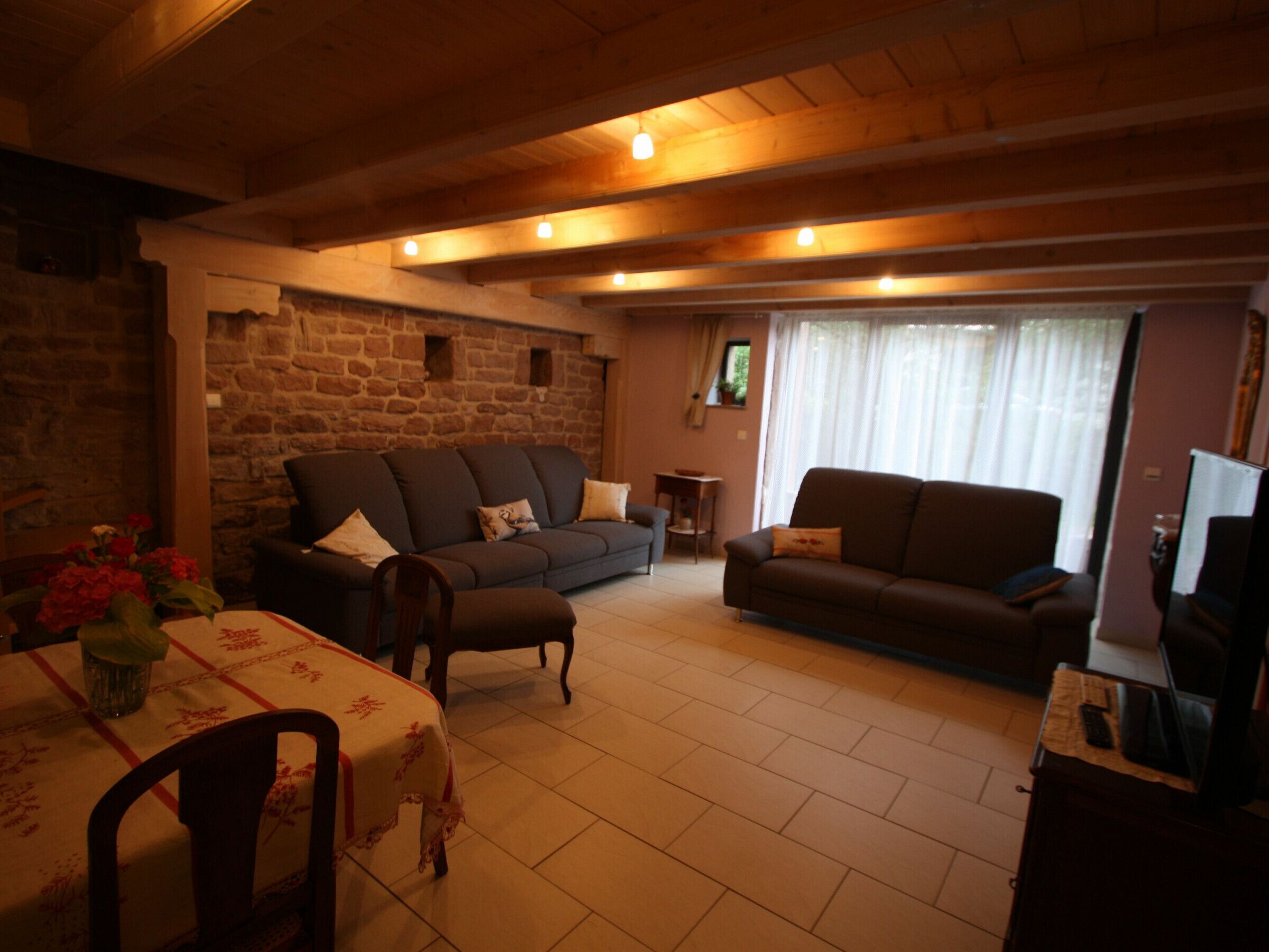 A cozy living room with a stone wall, two sofas, a dining table with a floral tablecloth, and a television on a wooden cabinet.