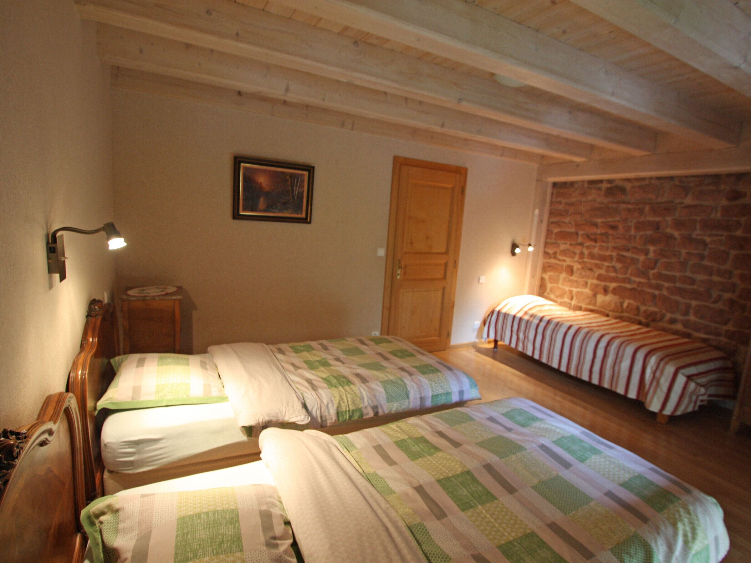 Une chambre avec deux lits simples, une tête de lit en bois, une petite table, une lampe fixée au mur, une porte et un mur en pierre.