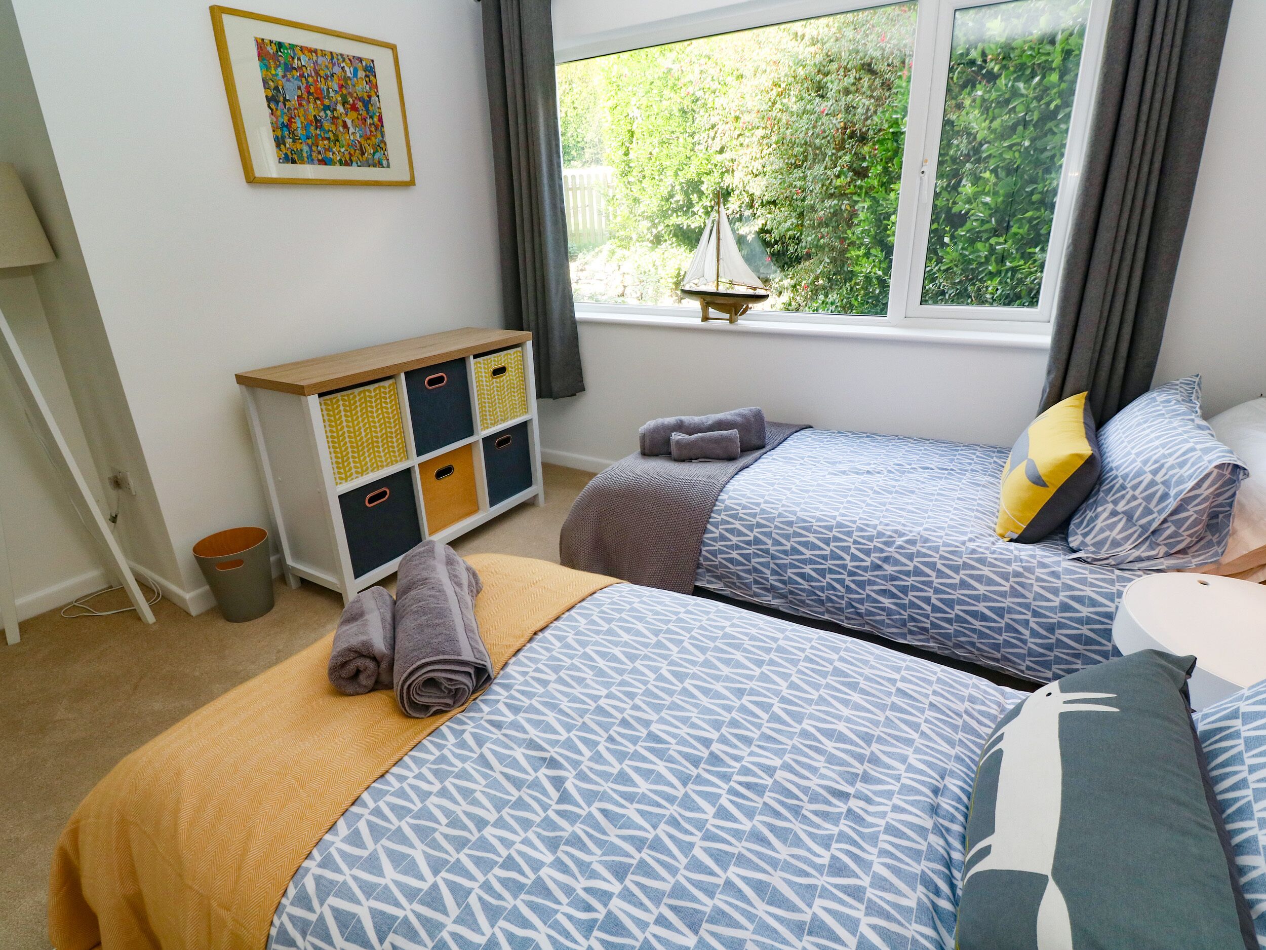 A bedroom with two beds, a window with a view of greenery, a bedside table, and a storage unit with multiple compartments.