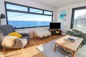 A modern living room with a sofa, a small table, a television, and a view of the sea.