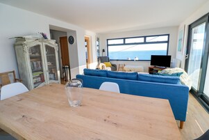 Una sala moderna con un ventanal grande con vistas al mar, un sofá azul, una mesa de comedor de madera y un aparador antiguo.