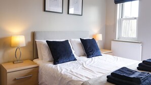 Un lit bien fait, avec une literie blanche et deux oreillers bleu foncé, une lampe de chevet et un tableau encadré au mur.